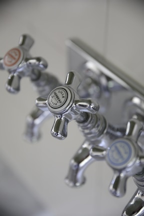 Three metallic cross-handled faucets are attached to a wall plate, each labeled with decorative text. The central faucet's label reads 'Lille Herbeau France'. The design is vintage with a polished chrome finish.