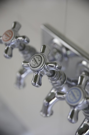 Three metallic cross-handled faucets are attached to a wall plate, each labeled with decorative text. The central faucet's label reads 'Lille Herbeau France'. The design is vintage with a polished chrome finish.