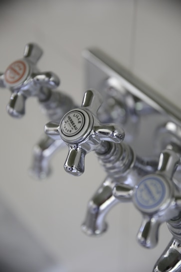 Three metallic cross-handled faucets are attached to a wall plate, each labeled with decorative text. The central faucet's label reads 'Lille Herbeau France'. The design is vintage with a polished chrome finish.