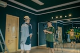 Two individuals in a room with dark walls, one is holding a camera on a stabilizer while the other stands facing the camera. There is a ladder leaning against the wall and a large mirror reflecting part of the scene. The room has a wooden door with some graffiti and studio lights on the ceiling.