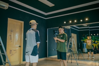 Two individuals in a room with dark walls, one is holding a camera on a stabilizer while the other stands facing the camera. There is a ladder leaning against the wall and a large mirror reflecting part of the scene. The room has a wooden door with some graffiti and studio lights on the ceiling.