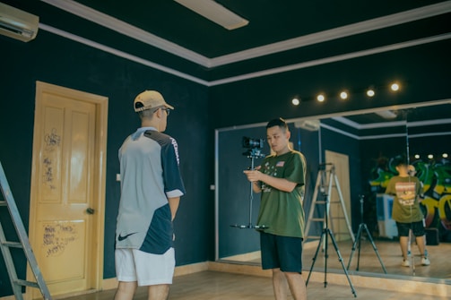 Two individuals in a room with dark walls, one is holding a camera on a stabilizer while the other stands facing the camera. There is a ladder leaning against the wall and a large mirror reflecting part of the scene. The room has a wooden door with some graffiti and studio lights on the ceiling.