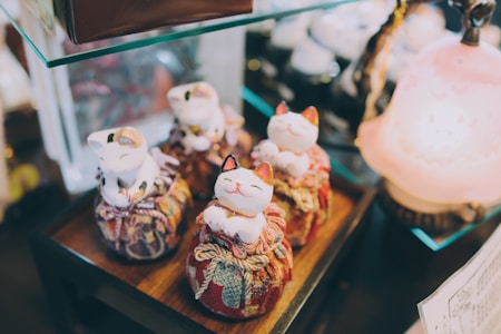 A set of four ceramic cat figurines are displayed on fabric cushions, each with a cheerful expression. The cats are arranged on a wooden surface, surrounded by a softly lit ambient setting, featuring delicate patterns on the cushions and a warm-toned lamp nearby.