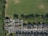 Aerial view of a residential neighborhood featuring rows of houses with gray roofs aligned along a street. A large green field borders the neighborhood, with a line of trees creating a visible boundary between the residential area and the open space. Several parked cars are seen along the street and in driveways, and shadows from the trees extend across the field.