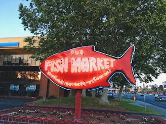 A large red neon fish-shaped sign indicates a fish market and seafood restaurant. In the background, there is a commercial building partially obscured by trees and a street with cars passing by. The sign stands on a brick base adorned with plants and greenery.