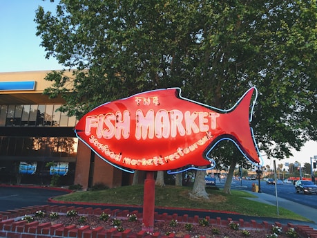 A large red neon fish-shaped sign indicates a fish market and seafood restaurant. In the background, there is a commercial building partially obscured by trees and a street with cars passing by. The sign stands on a brick base adorned with plants and greenery.