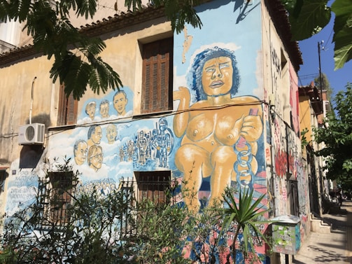 A mural painted on the side of a building depicts a large figure holding chains, with a group of diverse faces and scenes of protest or activism. The artwork is colorful and covers multiple walls, surrounded by greenery and an urban environment.