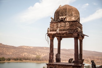 A group of monkeys is gathered on and around a historic stone pavilion with an ornate dome and columns, set against a backdrop of dry, rugged hills and a calm lake.