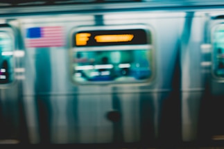 A motion-blurred subway train in transit with an American flag visible. The train's metallic exterior reflects light, and a digital display shows destination information in orange letters.