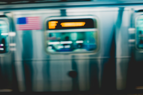 A motion-blurred subway train in transit with an American flag visible. The train's metallic exterior reflects light, and a digital display shows destination information in orange letters.