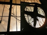 A large industrial fan, silhouetted, is positioned next to a barred window. The light filtering through the window creates a dramatic, shadowed effect, highlighting the geometric patterns of the fan and bars.