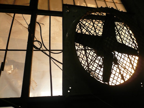 A large industrial fan, silhouetted, is positioned next to a barred window. The light filtering through the window creates a dramatic, shadowed effect, highlighting the geometric patterns of the fan and bars.