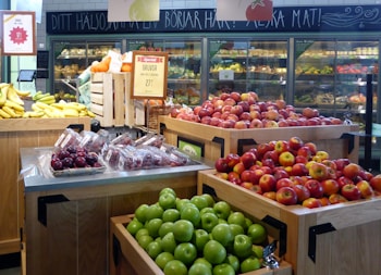 A grocery store produce section featuring displays of various fruits, including apples, bananas, plums, and grapes. Bright and colorful apples are placed prominently in wooden bins alongside bananas and green apples, with packaged plums and grapes visible. The background includes shelves stocked with a variety of fresh produce under a chalkboard with Swedish writing.