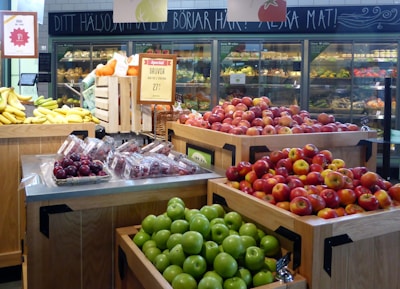 A grocery store produce section featuring displays of various fruits, including apples, bananas, plums, and grapes. Bright and colorful apples are placed prominently in wooden bins alongside bananas and green apples, with packaged plums and grapes visible. The background includes shelves stocked with a variety of fresh produce under a chalkboard with Swedish writing.