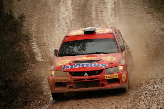 A rally car with bright red paint and multiple sponsor logos, including Mentos, races along a dirt track, kicking up a cloud of dust. The vehicle is equipped with racing modifications such as wide tires and a prominent air intake on the roof.