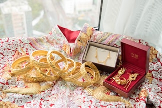An array of intricately designed gold jewelry, including bangles, a necklace, and earrings, are displayed on a richly embroidered fabric with red and gold patterns. Two jewelry boxes, one brown and the other red, hold some of the pieces, adding a touch of elegance to the presentation. The window in the background offers a blurred view of a cityscape.
