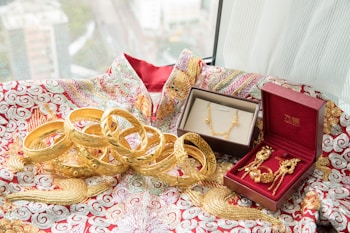 An array of intricately designed gold jewelry, including bangles, a necklace, and earrings, are displayed on a richly embroidered fabric with red and gold patterns. Two jewelry boxes, one brown and the other red, hold some of the pieces, adding a touch of elegance to the presentation. The window in the background offers a blurred view of a cityscape.