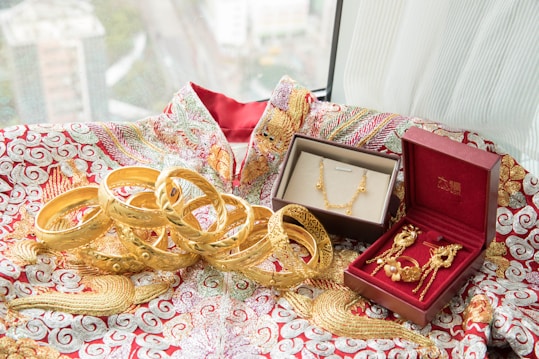 An array of intricately designed gold jewelry, including bangles, a necklace, and earrings, are displayed on a richly embroidered fabric with red and gold patterns. Two jewelry boxes, one brown and the other red, hold some of the pieces, adding a touch of elegance to the presentation. The window in the background offers a blurred view of a cityscape.