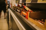 A close-up view of shelves in a store displaying various chocolate bars and snacks. The items are organized in cardboard boxes and there are prices listed above the shelves. The scene is slightly blurred in the background where a person is walking.
