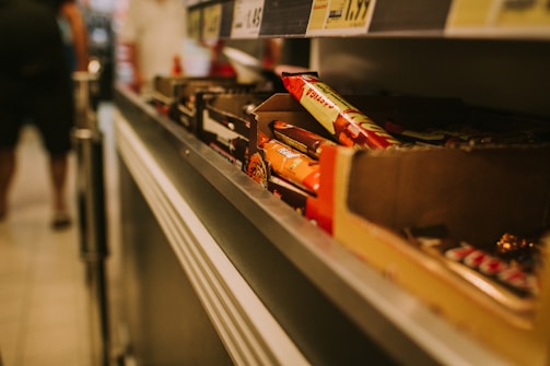 A close-up view of shelves in a store displaying various chocolate bars and snacks. The items are organized in cardboard boxes and there are prices listed above the shelves. The scene is slightly blurred in the background where a person is walking.