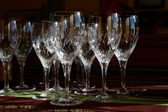 Several intricately designed crystal wine glasses are arranged on a table. The glasses catch and reflect light, creating sparkling highlights and intricate patterns. A dark, blurred background contrasts with colorful details in the foreground.