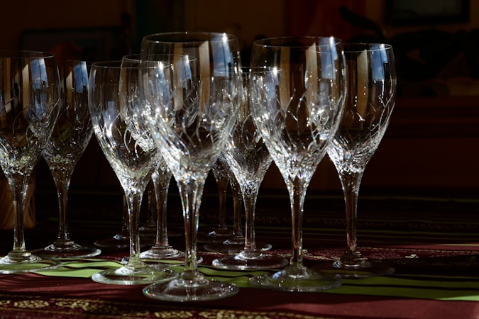 Several intricately designed crystal wine glasses are arranged on a table. The glasses catch and reflect light, creating sparkling highlights and intricate patterns. A dark, blurred background contrasts with colorful details in the foreground.