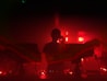 A person stands silhouetted against an intense red background, surrounded by music equipment with cables and lights suggesting a dynamic and energetic DJ set. The atmosphere is moody and vibrant, creating a sense of mystery and excitement.