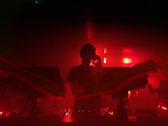 A person stands silhouetted against an intense red background, surrounded by music equipment with cables and lights suggesting a dynamic and energetic DJ set. The atmosphere is moody and vibrant, creating a sense of mystery and excitement.
