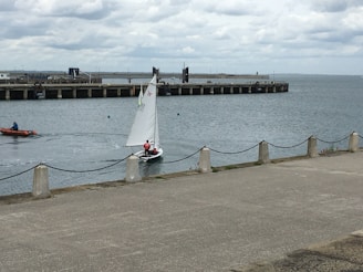 A small sailboat with two people aboard navigates calm waters near a pier. The boat is white with a single sail marked with symbols. In the background, a long concrete pier extends into the sea, lined with bollards. Another small motorized boat with a person in a blue jacket is moving to the left. The sky is overcast, with thick, grey clouds creating a muted atmosphere. A concrete walkway lines the water's edge, bordered by posts connected with chains.