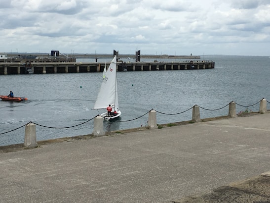A small sailboat with two people aboard navigates calm waters near a pier. The boat is white with a single sail marked with symbols. In the background, a long concrete pier extends into the sea, lined with bollards. Another small motorized boat with a person in a blue jacket is moving to the left. The sky is overcast, with thick, grey clouds creating a muted atmosphere. A concrete walkway lines the water's edge, bordered by posts connected with chains.