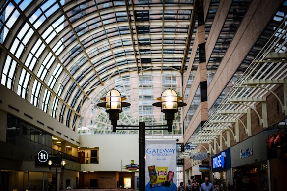 A spacious indoor walkway with a high, arched glass ceiling allows natural light to flood the area. Large hanging lanterns provide ambient lighting. There are various shops and signs visible, including a fitness center and a newstand advertisement. People can be seen walking through the area.