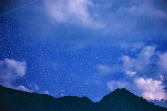A nighttime landscape featuring a silhouette of a mountain range against a star-filled sky. The sky is predominantly deep blue mixed with areas of lighter blue where the clouds are present.