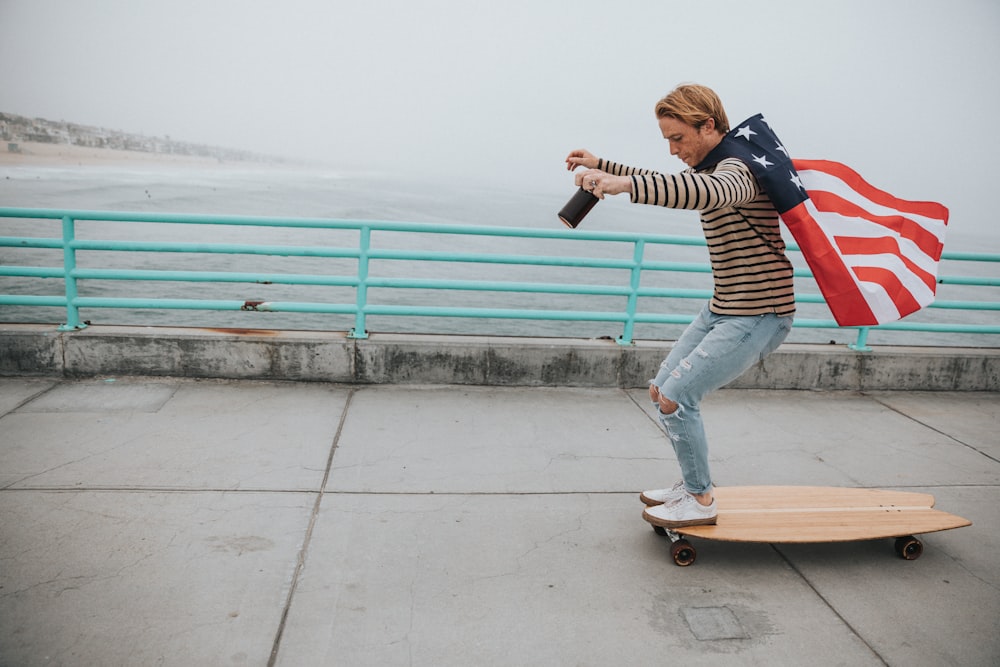 uomo che indossa un mantello con bandiera degli Stati Uniti mentre cavalca su un longboard vicino a uno specchio d'acqua durante il giorno