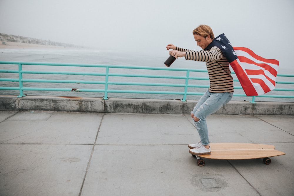 man wearing US flag cape while riding on longboard near body of water during daytime