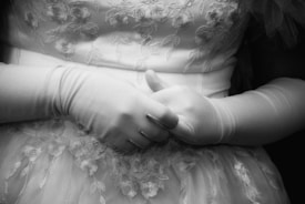 A close-up black-and-white photograph of a person wearing a floral-patterned dress and long gloves. The person's hands are gently clasped together, conveying a sense of formality and calmness.