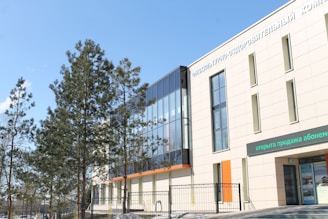 A modern building with large glass windows and a light-colored facade is visible. There are trees with green foliage in front of the structure, and the sky is clear with a few clouds. Text in another language is present on the building's facade and above a doorway.