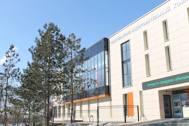 A modern building with large glass windows and a light-colored facade is visible. There are trees with green foliage in front of the structure, and the sky is clear with a few clouds. Text in another language is present on the building's facade and above a doorway.