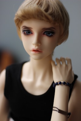 A realistic doll with short blonde hair and striking blue eyes wearing a black sleeveless top. The doll has a neutral expression and is posed with one hand touching the neck, adorned with a black beaded bracelet and a thin brown band.