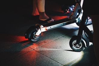 A close-up view of an electric scooter on a dimly lit pavement. The scooter's wheels and undercarriage are illuminated, casting red and white lights onto the ground. Partial views of people's legs are visible, indicating nighttime activity.