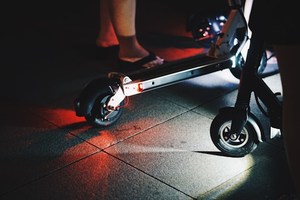 A close-up view of an electric scooter on a dimly lit pavement. The scooter's wheels and undercarriage are illuminated, casting red and white lights onto the ground. Partial views of people's legs are visible, indicating nighttime activity.