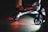 A close-up view of an electric scooter on a dimly lit pavement. The scooter's wheels and undercarriage are illuminated, casting red and white lights onto the ground. Partial views of people's legs are visible, indicating nighttime activity.