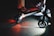 A close-up view of an electric scooter on a dimly lit pavement. The scooter's wheels and undercarriage are illuminated, casting red and white lights onto the ground. Partial views of people's legs are visible, indicating nighttime activity.