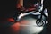 A close-up view of an electric scooter on a dimly lit pavement. The scooter's wheels and undercarriage are illuminated, casting red and white lights onto the ground. Partial views of people's legs are visible, indicating nighttime activity.