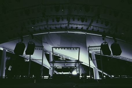 A large stage setup featuring an array of lighting equipment and large hanging speakers under an expansive canopy. The structure includes metal beams and pillars, illuminated with various lights focusing on the stage area.