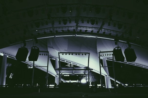 A large stage setup featuring an array of lighting equipment and large hanging speakers under an expansive canopy. The structure includes metal beams and pillars, illuminated with various lights focusing on the stage area.