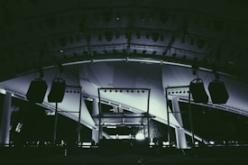 A large stage setup featuring an array of lighting equipment and large hanging speakers under an expansive canopy. The structure includes metal beams and pillars, illuminated with various lights focusing on the stage area.