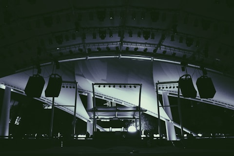 A large stage setup featuring an array of lighting equipment and large hanging speakers under an expansive canopy. The structure includes metal beams and pillars, illuminated with various lights focusing on the stage area.