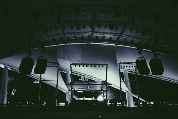 A large stage setup featuring an array of lighting equipment and large hanging speakers under an expansive canopy. The structure includes metal beams and pillars, illuminated with various lights focusing on the stage area.