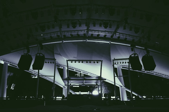 A large stage setup featuring an array of lighting equipment and large hanging speakers under an expansive canopy. The structure includes metal beams and pillars, illuminated with various lights focusing on the stage area.