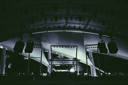 A large stage setup featuring an array of lighting equipment and large hanging speakers under an expansive canopy. The structure includes metal beams and pillars, illuminated with various lights focusing on the stage area.